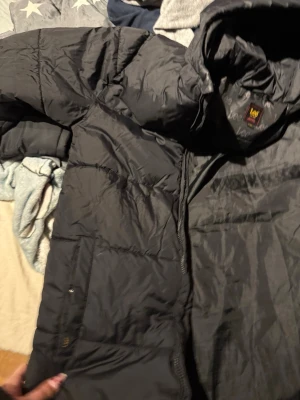 Svart dunjacka från Lee med huva - Säljer en svart dunjacka från Lee med huva och dragkedja. Jackan har två sidofickor med dragkedja och diskret Lee-logga på bröstet. Perfekt för kalla vinterdagar och riktigt skön att bära. Klassisk quiltad design. aldrig använd