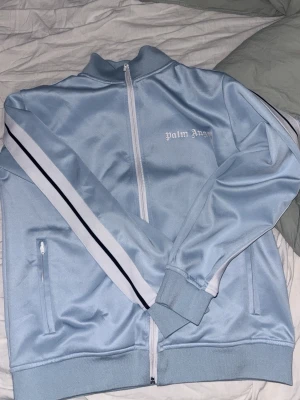 Ljusblå ziptröja från Palm Angels - Säljer en ljusblå ziptröja från Palm Angels med vita och svarta ränder längs ärmarna. Tröjan har hög krage, dragkedja framtill och två fickor med dragkedja. Palm Angels-logga broderad i vitt på bröstet. Perfekt för en sportig och trendig look.