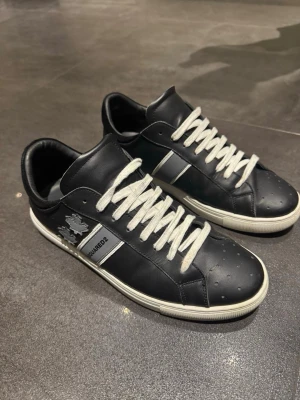 Svarta Dsquared2 sneakers i läder - Snygga svarta sneakers från Dsquared2 i äkta läder med vita kontrastsnören och vit sula. Skorna har perforerade detaljer på tån, silverfärgad logga på sidan och ikonisk lönnlöv-design. Perfekt för dig som vill ha en clean och stilren look.