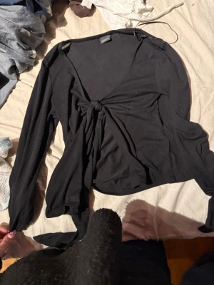 Svart omlottopp med knyt - Snygg svart omlottopp med knyt framtill och långa ärmar. Toppen har en djup v-ringning och är tillverkad i ett mjukt, stretchigt material. Perfekt att styla med jeans eller kjol för en trendig look.