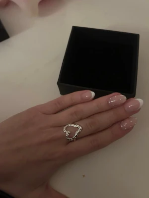 Silvrig ring med hjärtformad design - Unik silvrig ring med en öppen hjärtformad design. Ringen har en organiskt vriden struktur som ger ett trendigt och modernt intryck. Perfekt accessoar för dig som gillar stilrena men ändå iögonfallande smycken. Levereras i svart ask.