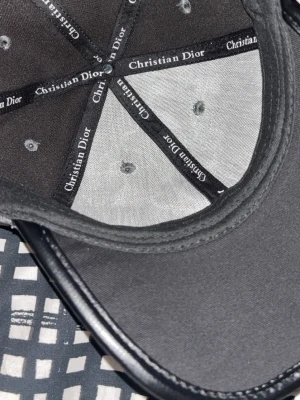 Grå mönstrad keps från Christian Dior - Snygg keps från Christian Dior i grått med ett stilrent monogrammönster över hela kepsen. Kepsen har svarta detaljer och logotyp på insidan. Materialet ser ut att vara en blandning av textil och syntet, vilket ger en exklusiv känsla. 