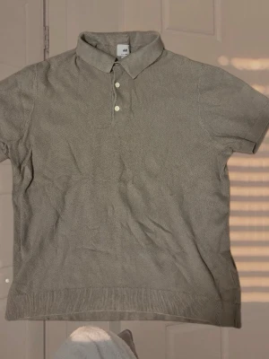 Grå pikétröja från H&M - Klassisk grå pikétröja från H&M med korta ärmar och tre knappar framtill. Tröjan har en ribbad kant nedtill och vid ärmsluten samt en mjuk krage. Perfekt för en clean och enkel stil.