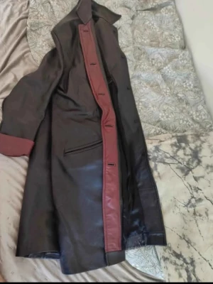 Lång svart skinnjacka med vinröda detaljer - Snygg lång svart skinnjacka med vinröda detaljer längs knappraden och vid ärmsluten. Jackan har bred krage och raka fickor framtill. Perfekt för dig som vill sticka ut med en unik och stilren look. Jackan är också hemmagjord och är otroligt välgjord!