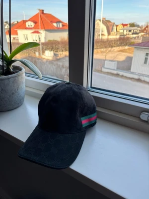 Gucci keps - Svart keps från Gucci med diskret GG-mönster i tyget och klassiska gröna och röda ränder på sidan. Böjd skärm och justerbar passform. Snygg accessoar som lyfter din outfit med lyxig känsla.