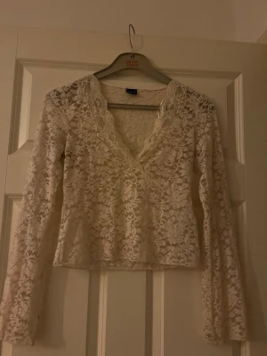 Vit spetsblus från Gina Tricot - Superfin vit blus i spets från Gina Tricot med v-ringning och långa ärmar. Blusen har ett blommigt spetsmönster och är lätt transparent, perfekt för att skapa en romantisk look. Snygg att styla med både jeans och kjol.