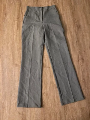 Grå kostymbyxor med pressveck - Snygga grå kostymbyxor med klassiska pressveck och rak passform. Byxorna har hög midja och stängs med dragkedja och knapp framtill. Perfekta för en stilren look. 