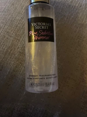 Victoria's Secret Pure Seduction Shimmer - En shimmer body mist från Victoria's Secret i doften Pure Seduction Shimmer. Flaskan är genomskinlig med svart etikett och rosa text, har skimrande partiklar som ger huden en glittrig finish. Perfekt för dig som vill dofta gott och glänsa lite extra.