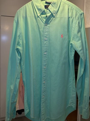 Grön skjorta från Polo Ralph Lauren ny skick - Snygg grön skjorta från Polo Ralph Lauren i slim fit-modell. Skjortan har klassisk krage, långa ärmar och knappar framtill. Broderad rosa logga på bröstet ger en stilren touch. Perfekt till jeans eller chinos för en clean look.