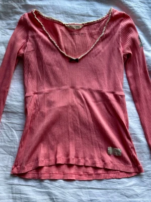Rosa ribbad långärmad topp Odd Molly - Supersöt rosa ribbad topp från Odd Molly med lång ärm och v-ringad hals. Dekorerad med vit spetskant och liten rosett framtill. Tillverkad i mjuk bomull som känns skön mot huden. Perfekt för dig som gillar en feminin och avslappnad stil.