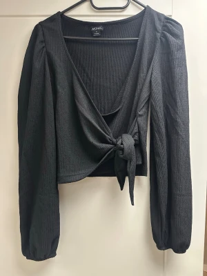 Svart omlottopp från Monki S - Säljer en svart omlottopp från Monki med långa puffärmar och knyt framtill. Toppen är ribbad och croppad, vilket ger en trendig look. Perfekt för dig som gillar stilrena och moderna plagg med snygga detaljer.