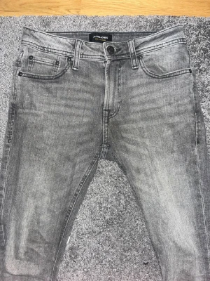 Grå slim fit jeans från Jack & Jones - Snygga grå slim fit jeans från Jack & Jones med klassisk femficksmodell och svarta detaljer vid fickorna. Jeansen har en tvättad look och är tillverkade i mjuk denim som ger en skön passform. Perfekta för en avslappnad och trendig stil.