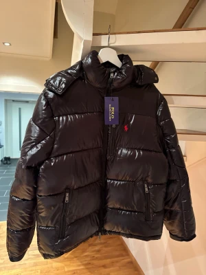 Svart dunjacka från Polo Ralph Lauren - Snygg svart dunjacka från Polo Ralph Lauren med blank finish och röd logga på bröstet. Jackan har huva, två framfickor med dragkedja och hög krage. Perfekt för kalla vinterdagar och riktigt trendig look.