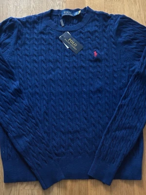 Mörkblå kabelstickad tröja Polo Ralph Lauren - Snygg mörkblå kabelstickad tröja från Polo Ralph Lauren med rund halsringning och röd broderad logga på bröstet. Tröjan har långa ärmar och ribbade muddar. Perfekt för dig som vill ha en klassisk och stilren look.