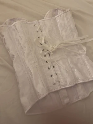 Vit brokadkorsett med snörning - Säljer en snygg vit korsett i glansigt brokadtyg med vackert mönster. Framsidan har snörning med satinband och baksidan har dragkedja för enkel påklädning. Ger en klassisk siluett och passar perfekt till olika outfits.