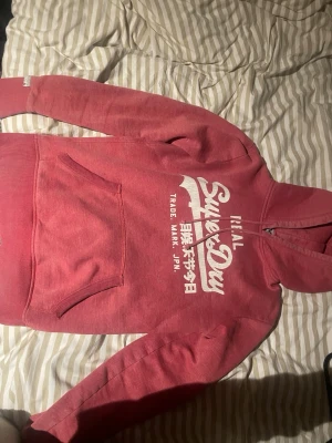 Rosa/röd hoodie från Superdry med tryck - Säljer en snygg rosa/röd hoodie från Superdry med vitt tryck på bröstet och japanska tecken. Hoodien har en stor känguruficka framtill, ribbade muddar och en klassisk huva. Perfekt för dig som gillar streetwear och vill ha något bekvämt och stilrent.