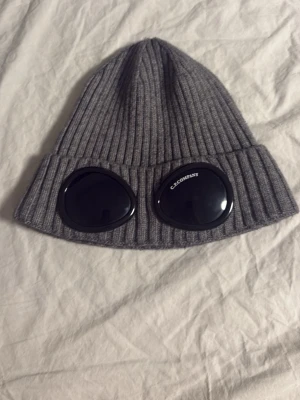 Grå C.P.COMPANY mössa - Snygg grå ribbstickad mössa med uppvikt kant. Klassisk modell som passar perfekt till kalla dagar. Tillverkad i mjukt och stretchigt material som sitter skönt på huvudet. Enkel att matcha med olika outfits.