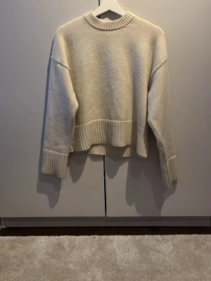 Beige stickad tröja från H&M - Mysig beige stickad tröja från H&M med bred ribbad mudd vid ärmslut, nederkant och halsringning. Tröjan har en avslappnad passform och är tillverkad i mjukt material som passar perfekt till kyliga dagar.