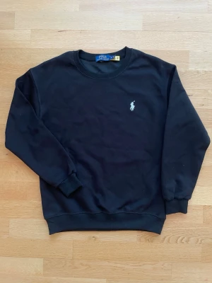 Svart sweatshirt från Polo Ralph Lauren - Svart sweatshirt från Polo Ralph Lauren med rund hals och ribbade muddar. Tröjan har den klassiska vita broderade loggan på bröstet och är gjord i mjukt bomullstyg. Enkel och stilren modell som funkar till allt.