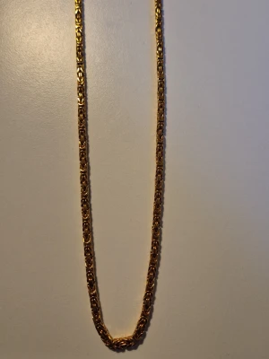 Guldhalsband från kejsar.se - Snyggt halsband i guldfärg med en robust, flätad kedjedesign. Halsbandet har en glänsande finish och känns rejält, perfekt för dig som gillar statement-smycken. Materialet ser ut som metall med guldfinish. Vikten är 58 gram enligt vågen på bilden.