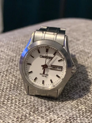 Champion silverfärgad klocka - Stilren klocka från Champion med vit urtavla och tydliga markeringar. Boetten och länken är i silverfärgad metall och klockan har dag- och datumvisning. Saphirglas och vattentät till 100 meter. Perfekt accessoar för dig som gillar klassisk design. Funkar som ska och nyligen byt batteri 