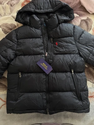 Svart pufferjacka Polo Ralph Lauren - Snygg svart pufferjacka från Polo Ralph Lauren med huva och dragkedja framtill. Jackan har två sidofickor med dragkedja och den klassiska röda loggan broderad på bröstet. Perfekt för kalla vinterdagar och riktigt skön att bära.