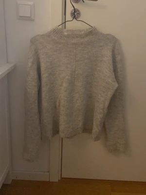 Ljusgrå stickad tröja med rund hals - Mysig ljusgrå stickad tröja med rund hals och långa ärmar. Tröjan har en enkel, rak passform och ribbstickade muddar vid hals, ärmslut och nederkant. Perfekt för kalla dagar och enkel att matcha med olika outfits.