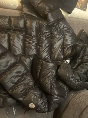 Moncler pufferjacka - Svart pufferjacka från Moncler med glansig finish, huva och dragkedja framtill. Jackan har Moncler-logga på ärmen, praktiska fickor med dragkedja och coolt serietidningsmönster på insidan. Perfekt för dig som vill ha en snygg och varm jacka med street vibe. Pris kan diskuteras vid snabb affär!