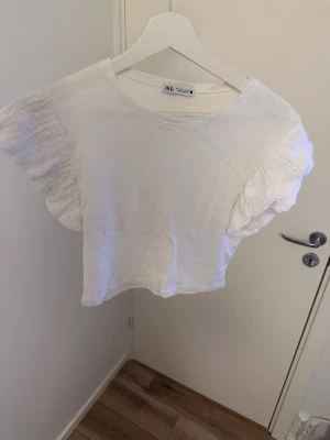 Vit croppad topp med volangärm Zara - Superfin vit croppad topp från Zara med korta volangärmar. Toppen har en enkel rund halsringning och är tillverkad i ett mjukt material som känns skönt mot huden. Perfekt att styla med jeans eller kjol för en trendig look.