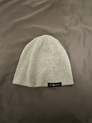 Ljusgrå mössa från Sail Racing - Säljer en stilren ljusgrå mössa från Sail Racing. Mössan är ribbstickad i mjukt material och har en diskret svart logolapp framtill. Perfekt för att hålla värmen och samtidigt se snygg ut under kalla dagar. Hör av er vid frågor!