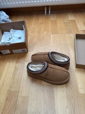 Bruna UGG tofflor med fårull - Säljer ett par bruna UGG tofflor i mocka med mjukt vitt fårullsfoder. Tofflorna har en dekorativ kant i svart och vitt mönster runt öppningen och UGG-logga på sidan. Perfekta för att hålla fötterna varma hemma.