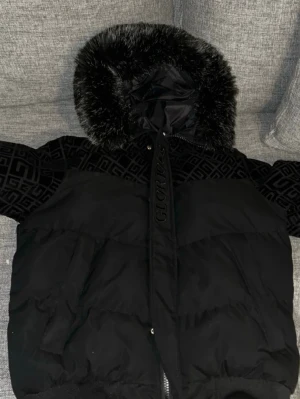 Svart pufferjacka från glorious gangsta - Snygg svart pufferjacka från Gicri med mönstrade detaljer upptill och på huvan. Jackan har en stor huva med svart fuskpäls och dragkedja framtill. Materialet är en mix av glansig och mönstrad textil som ger en lyxig känsla.