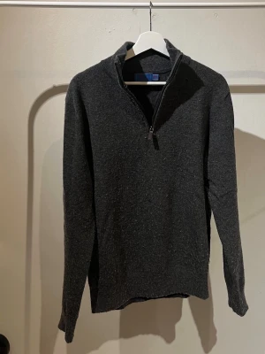 Halfzip Tröja  - Mörkgrå stickad tröja med halv dragkedja och hög krage. Tröjan har lång ärm och ribbade muddar vid ärmslut och nederkant. Perfekt för lager på lager och en clean look.