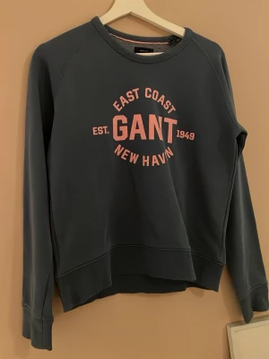 Blå sweatshirt från GANT - Mysig blå sweatshirt från GANT med tryck i rosa framtill. Tröjan har rund halsringning, långa ärmar och ribbade muddar vid ärmslut och nederkant. Perfekt för en avslappnad och stilren look.