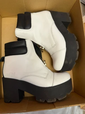 Chunky vita stövletter från Johnny bulls nypris 1300 - Säljer ett par chunky vita stövletter  med grov svart platåsula och dragkedja framtill med ringdetalj. Skorna har svart vadderad kant upptill och är tillverkade i skinn. Perfekta för dig som gillar statement boots och vill ha något som sticker ut.