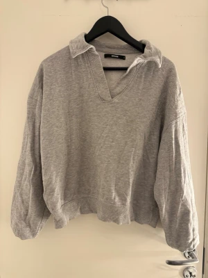 Grå collegetröja från Bik Bok - En oversized grå collegetröja från Bik Bok med krage och v-ringad hals. Tröjan har långa ärmar och ribbade muddar vid ärmslut och nederkant. Perfekt för en avslappnad och trendig look. 