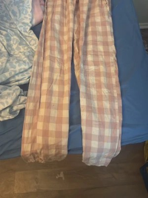 Rutiga pyjamasbyxor i rosa och vitt - Mysiga pyjamasbyxor med rutigt mönster i ljusrosa och vitt. Byxorna har rak passform och resår i midjan för extra komfort. Perfekta för slappa dagar hemma eller när du vill ha det extra bekvämt på natten.