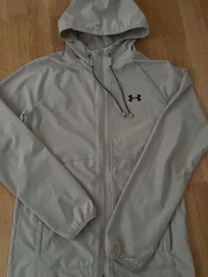Ljusgrå vindjacka från Under Armour - Snygg ljusgrå vindjacka från Under Armour med huva och hel dragkedja framtill. Jackan har justerbar dragsko i huvan, två sidofickor och elastiska muddar vid ärmsluten. Perfekt för blåsiga dagar och sportiga outfits.