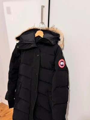 Svart dunjacka från Canada Goose - Säljer en svart dunjacka från Canada Goose med klassisk patch på ärmen och stor huva med fuskpäls. Jackan är quiltad, har dragkedja och knappar framtill samt långa ärmar. Perfekt för kalla vinterdagar och riktigt snygg streetstil.