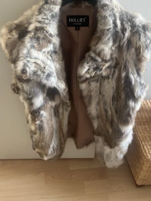Pälsväst i naturfärger från Hollies - Snygg pälsväst från Hollies i storlek M. Västen har ett fluffigt och lyxigt utseende med naturfärgad fuskpäls i toner av beige, grått och vitt. Perfekt att slänga över en hoodie eller tröja för en trendig lager-på-lager-look.