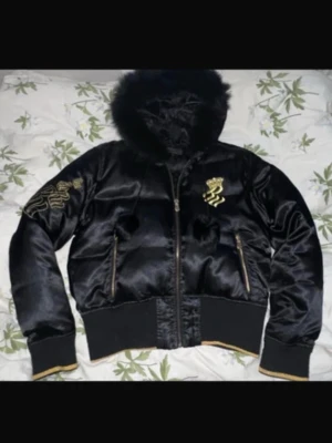 Svart pufferjacka med gulddetaljer - Snygg svart pufferjacka med glansig finish och stora guldbroderade ormar på bröstet och ryggen. Jackan har en stor huva med fuskpäls, ribbade muddar och dragkedjor i guld. Perfekt för kalla vinterdagar och riktigt statement-plagg.