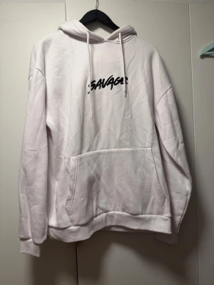 Vit hoodie från Black Squad - Säljer en oversized vit hoodie från Black Squad med texten 'SAVAGE' i svart på bröstet. Hoodien har stor känguruficka framtill, dragsko i huvan och mjukt material på insidan. Perfekt för en chill och avslappnad stil.