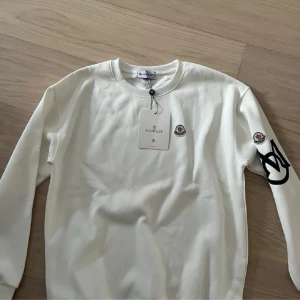 Vit sweatshirt från Moncler - Snygg vit sweatshirt från Moncler med rund halsringning och Moncler-logga på bröstet samt ärmen. Tröjan har ett svart tryck på ena ärmen och är tillverkad i mjukt material. Perfekt för en clean och stilren look.