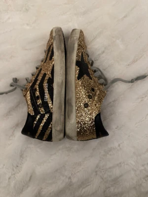 Guldglittriga sneakers från Golden Goose - Unika sneakers från Golden Goose med guldglittrig ovandel, svarta detaljer och stjärna på sidan. Skorna har grå skosnören och vit sula. Märket syns på plösen och insidan. Perfekta för dig som vill sticka ut med coola detaljer och trendig look.