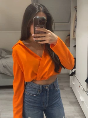 Orange croppad hoodie med huva - Snygg orange croppad hoodie med huva och omlott-design framtill. Hoodien har långa ärmar och en avslappnad passform, perfekt att matcha med höga jeans för en trendig look.