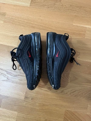Svarta Nike Air Max 97 sneakers - Snygga svarta Nike Air Max 97 sneakers med röda detaljer och ikonisk vågig design. Skorna har en synlig Air-sula längs hela foten och är tillverkade i en mix av textil och syntetmaterial. Perfekta för dig som gillar streetwear och sportig stil.