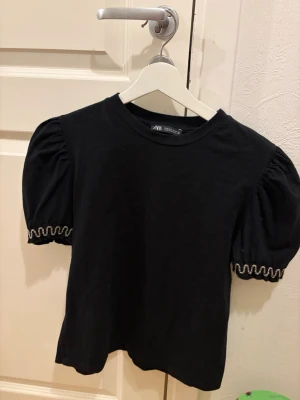 Svart topp med puffärm från Zara - Svart topp från Zara med puffärmar och rund halsringning. Ärmarna har en snygg volangkant med gulddetaljer som ger en unik touch. Tillverkad i mjuk bomull som känns skön mot huden. Perfekt för dig som vill ha en enkel men ändå trendig topp.