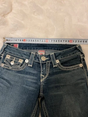 Blå True Religion jeans med kontrastsömmar - Säljer ett par blå jeans från True Religion med tydliga vita kontrastsömmar och klassiska fem fickor. Jeansen har låg midja, raka ben och coola detaljer som logga på knappen och ficklock med knapp. Perfekta för en avslappnad och trendig stil.