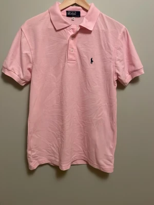 Ljusrosa pikétröja från Ralph Lauren - Klassisk ljusrosa pikétröja från Polo Ralph Lauren med korta ärmar och krage. Tröjan har två knappar vid halsen och den ikoniska broderade loggan i svart på bröstet. Tillverkad i 100% mjuk bomull som känns skön mot huden. Perfekt för en avslappnad men stilren look.