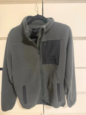 Grå fleecejacka från Everest - Mysig grå fleecejacka från Everest med hel dragkedja, huva och tre fickor med svarta detaljer. Bröstficka med dragkedja och broderad logga. Perfekt för lager-på-lager och outdoor. Tillverkad i mjuk fleece som håller värmen.